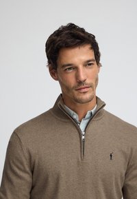 Brun zip-up tröja i mjukt material med hög krage och en liten logotyp på vänstra sidan. Lager-på-lager över en ljusblå skjorta med knappar.