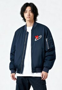 Giacca bomber blu navy con zip frontale, polsini a coste e ricamo di un cuore rosso con spilla da balia sul petto sinistro. Tessuto dalla texture liscia.