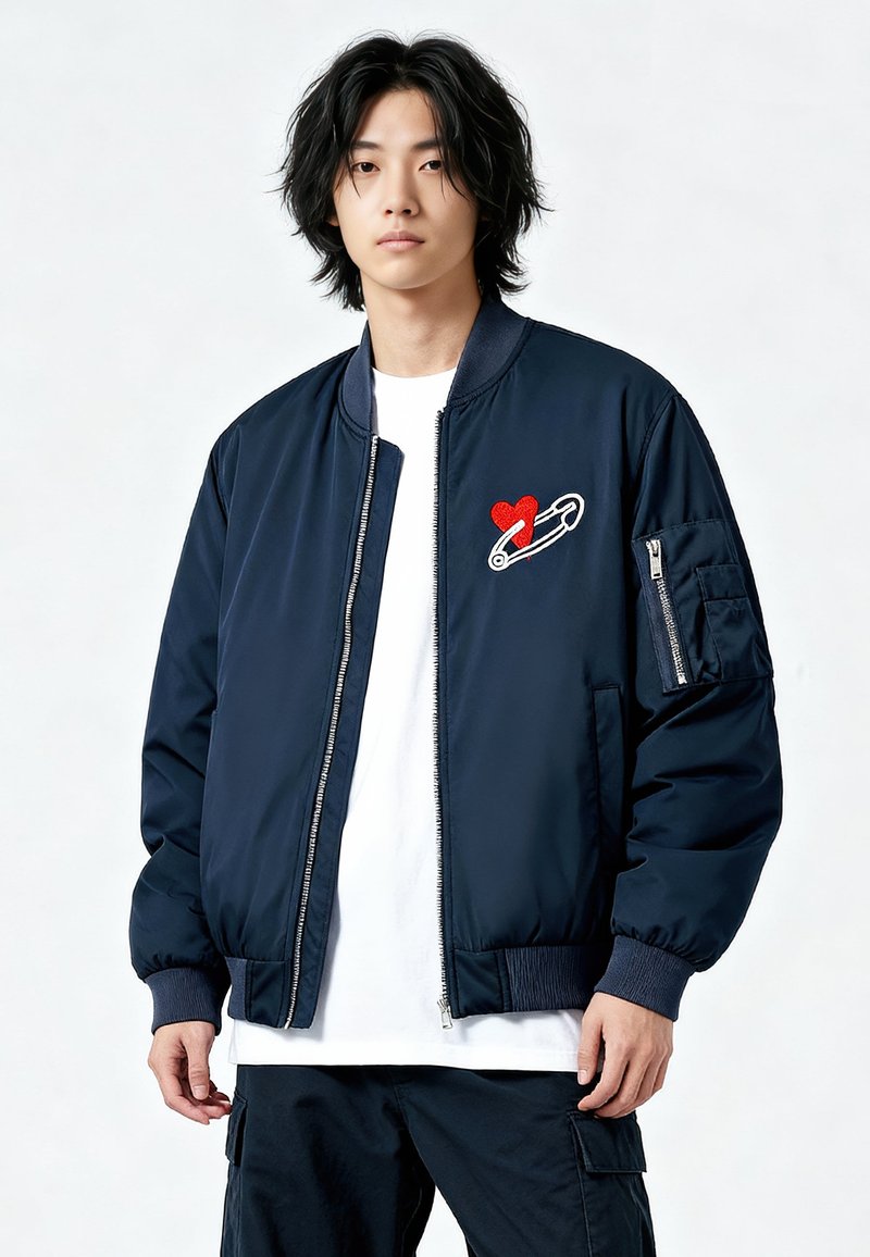 Giacca bomber blu navy con zip frontale, polsini a coste e ricamo di un cuore rosso con spilla da balia sul petto sinistro. Tessuto dalla texture liscia.