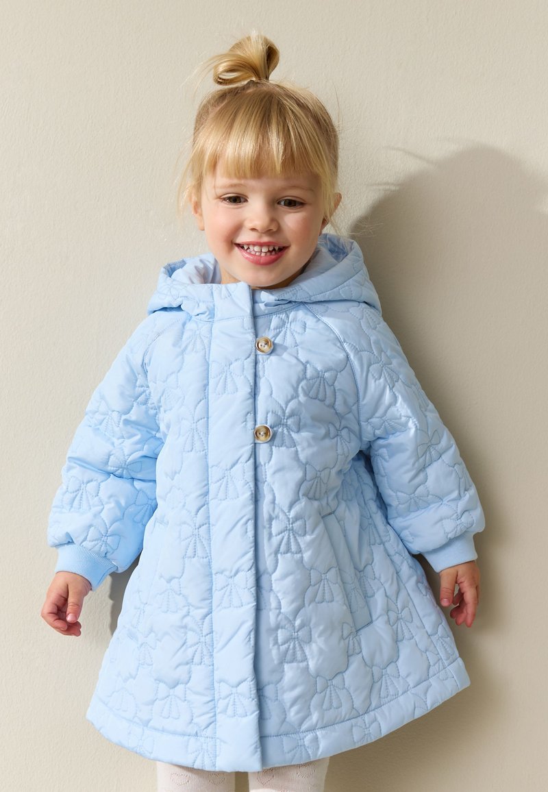 Jeune enfant souriant aux cheveux blonds attachés en chignon, portant un manteau matelassé bleu clair avec une broderie florale et des boutons en bois.