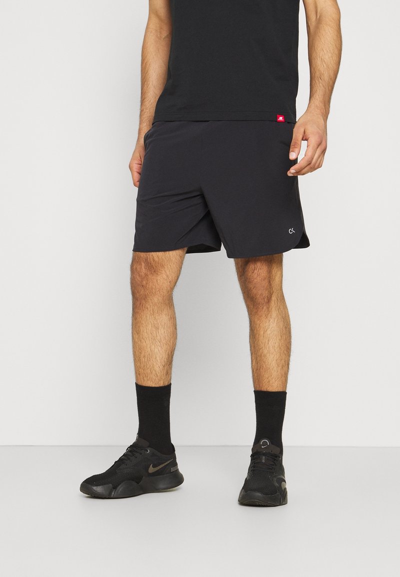 Homme portant un short de sport noir, des chaussettes noires, des baskets noires et un t-shirt noir, debout devant un fond blanc.