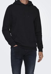 Sudadera negra hecha de tela suave, con bolsillo tipo canguro, capucha con cordón y puños acanalados. Combinada con vaqueros oscuros.