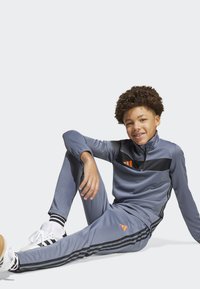 Chándal atlético gris con acentos negros y logo naranja. Presenta un cuello con cremallera y pantalones ajustados con franjas laterales. Zapatos blancos.