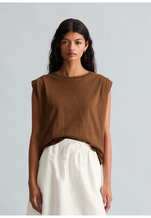 Kobieta o długich ciemnych włosach, ubrana w brązową bluzkę bez rękawów i białą spódnicę z wysokim stanem ze sznurkiem w talii, na jednolitym szarym tle.