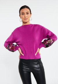 Maglione fucsia con un classico collo a giro e dettagli a motivo con paillettes sulle maniche. Presenta polsini e orlo a costine. Indossato con pantaloni neri.