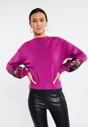 Pull fuchsia à col ras du cou classique avec des accents en sequins sur les manches. Il présente des poignets et un ourlet côtelés. Porté avec un pantalon noir.