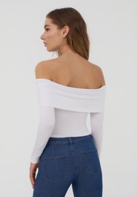 Body bianco a spalle scoperte con maniche lunghe e design aderente, indossato con jeans blu scuro a vita alta. Texture liscia, dettagli minimi.