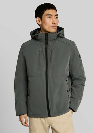 Man draagt een grijze capuchonjas met rits en beige broek, staat met ontspannen handen, kijkt naar de zijkant tegen een effen achtergrond.