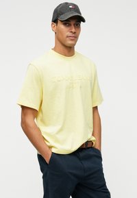 Lys gul bomulls-T-skjorte med preget "TOMMY JEANS"-logo. Kombinert med marineblå bukser og en mørk caps med logoakkenter.