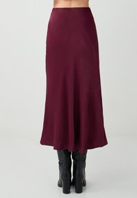 Donna che indossa una gonna bordeaux alta in vita, lunga fino alla caviglia, abbinata a stivali neri con tacco al ginocchio, su uno sfondo semplice.