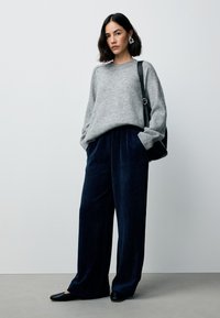 Maglione grigio a maglia con maniche larghe, abbinato a pantaloni a zampa d'elefante in velluto a coste blu scuro. Scarpe nere basse e una borsa a tracolla nera completano il look.