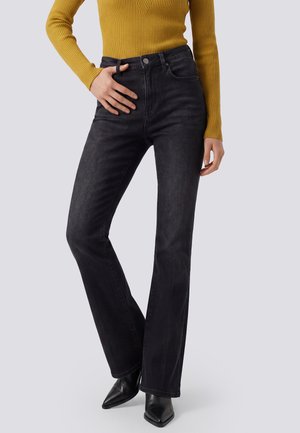 Modelo con vaqueros negros acampanados, un jersey de canalé color mostaza y botas negras de punta, posando ante un fondo de estudio gris claro.