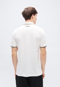 Λευκό πόλο μπλουζάκι με μαύρο γιακά και λεπτομέρειες στα μανίκια. Στη ράχη βρίσκεται το λογότυπο "BOSS" κοντά στον λαιμό. Ομαλή υφή ύφασματος.