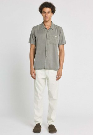 Homme debout portant une chemise grise à manches courtes boutonnée, un pantalon blanc et des chaussures marron foncé sans lacets sur un fond uni.