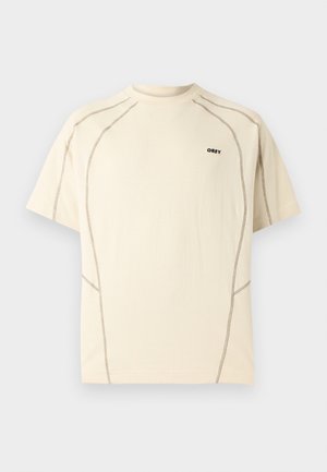 Beige kortermet T-skjorte med mørk kontrastsøm og liten "OBEY"-logo på venstre bryst mot lys bakgrunn.