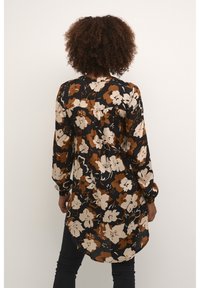 Femme aux cheveux bouclés portant une tunique noire à manches longues avec un motif floral marron et beige, associée à un pantalon noir, vue de dos.