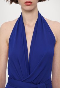 Niebieska sukienka na szyję z głębokim dekoltem w kształcie litery V i marszczonym materiałem. Posiada wiązany pasek w talii dla lepszego dopasowania oraz gładką fakturę.