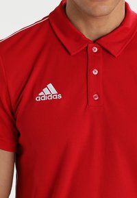 Rotes Poloshirt mit einem Kragen, drei Knöpfen und weißen Streifen auf den Schultern. Mit einem gestickten weißen Adidas-Logo.