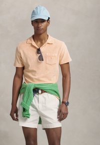 Lichtoranje polo met korte mouwen en een zak, gestyled met een groene trui om de taille, witte korte broek en een lichtblauwe pet.