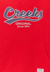 Felpa rossa con cappuccio e tasca a marsupio; logo "Creeks" in grigio scuro e la scritta "ORIGINAL Since 1974" stampata sotto in bianco. Tessuto morbido.
