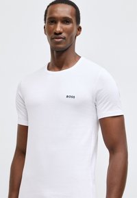 Bílé bavlněné tričko s kulatým výstřihem, krátkými rukávy a malým černým logem "BOSS" na levé straně hrudníku.