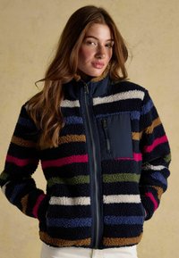 Fleece jacket featuring bold multicolored stripes in navy, pink, green, and brown. Zipper closure and a chest pocket with zip. Soft texture.