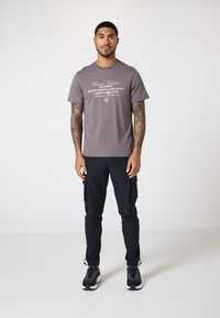 Graues Baumwoll-T-Shirt mit Frontschriftzug, kombiniert mit schwarzen Cargo-Hosen mit Seitentaschen und schwarzen Sneakers mit weißen Sohlen.
