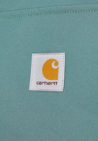 Teal stof med en tekstureret overflade har et Carhartt-mærke, hvid baggrund med et fremtrædende gult logo og rød tekst nedenfor.