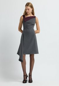 Graues ärmelloses Kleid mit einzigartigem kastanienbraunem geometrischem Ausschnitt, asymmetrischem Saum und subtiler Textur, kombiniert mit schwarzen Schuhen.