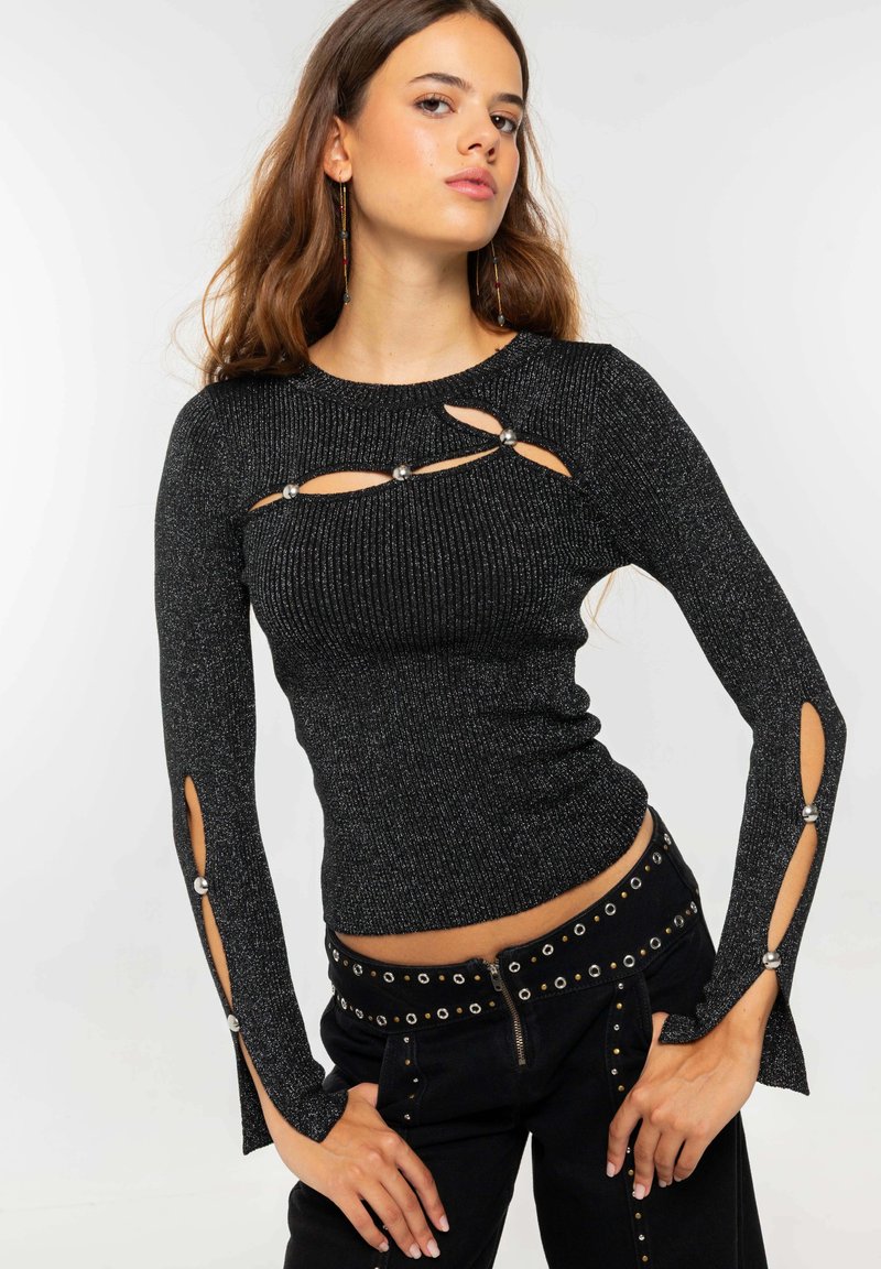 Top negro de manga larga con detalles recortados y acentos metálicos. La tela tiene un ligero brillo. Combinado con pantalones negros de cintura alta con tachuelas.