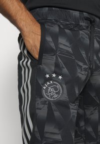 Pantalons de sport noirs avec un motif géométrique, présentant un logo rond d'Ajax Amsterdam et des bandes argentées accentuées sur les côtés.