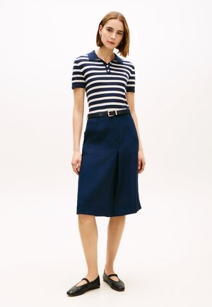 Kobieta ubrana w granatowo-białą paskowaną koszulkę polo z krótkim rękawem, granatową spódnicę do kolan z kieszeniami, czarny pasek i czarne baleriny typu Mary Jane, stojąca.