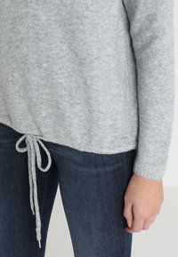 Lichtgrijze, getextureerde trui met een koord aan de onderkant. Lange mouwen en een relaxte pasvorm, gecombineerd met donkerblauwe jeans.