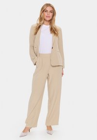 Blazer beige sur mesure avec un bouton, assorti à un pantalon large beige. Le mannequin porte un t-shirt blanc et des talons avec des accents argentés.