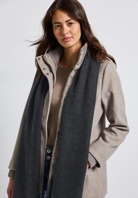 Grå tørklæde draperet over en beige frakke med ribbet kravestruktur, der har lynlås og trykknapper, båret over en lys bluse og denim.