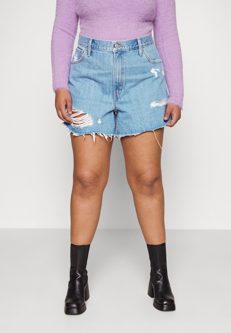 Personne portant un haut à manches longues violet en tissu duveteux, une mini-jupe en denim bleu délavé et des bottines à talons noires, debout sur un sol gris.