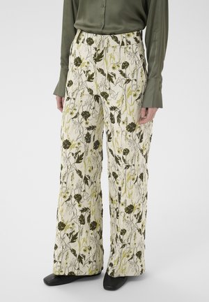 Hlače širokih hlačnic z osnobo v barvi slonokoščene kosti in zelenim cvetličnim vzorcem, nošene z dolgo zeleno srajco in črnimi čevlji.