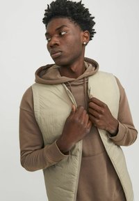 Gilet matelassé beige sur un sweat à capuche marron, avec une fermeture éclair et des poches latérales, au toucher lisse et à coupe décontractée.