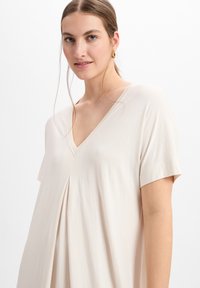 Beige V-Ausschnitt-Oberteil mit kurzen Ärmeln und Faltendetail vorne, aus einem weichen, leichten Stoff mit glatter Textur gefertigt.