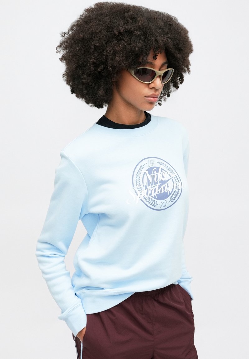 Sweatshirt bleu clair avec un logo rond en blanc et bleu marine. Présente des poignets et un col côtelés, associé à un short noir et des lunettes de soleil vertes.