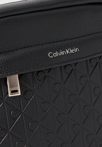 Sac à main en cuir noir texturé, présentant un motif géométrique avec des triangles en relief, un fermoir en métal et un logo "Calvin Klein" en argent.
