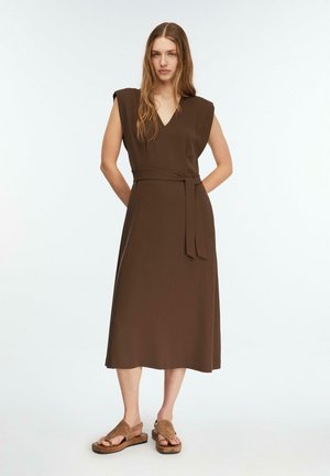 Kobieta stojąca z rękami za plecami, ubrana w brązową, bezrękawną midi sukienkę z paskiem oraz brązowe sandały na jednolitym, jasnym tle.