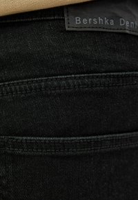 Mustat farkut, joissa on sileä pinta, erottuvat ompeleet ja näkyvä merkkilogo, sijoitettuna vyötärön ylle.