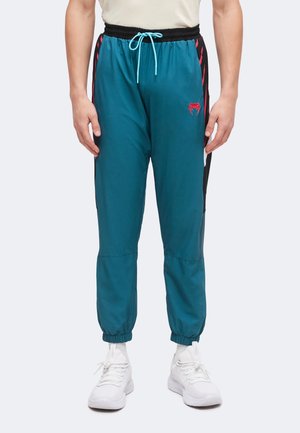 Mand iført turkis joggingbukser med sorte og røde sidelommer, sort elastisk linning med blå snor og hvide sneakers.