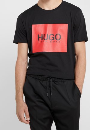 Mann trägt ein schwarzes T-Shirt mit einem roten Quadrat und dem Schriftzug „HUGO HUGO BOSS“, kombiniert mit schwarzen Hosen mit Kordelzug.