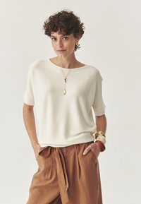Letvægts, cremefarvet strikket sweater med en bred halsudskæring og korte ærmer, parret med brune, højtaljede bukser som er bundet i taljen.