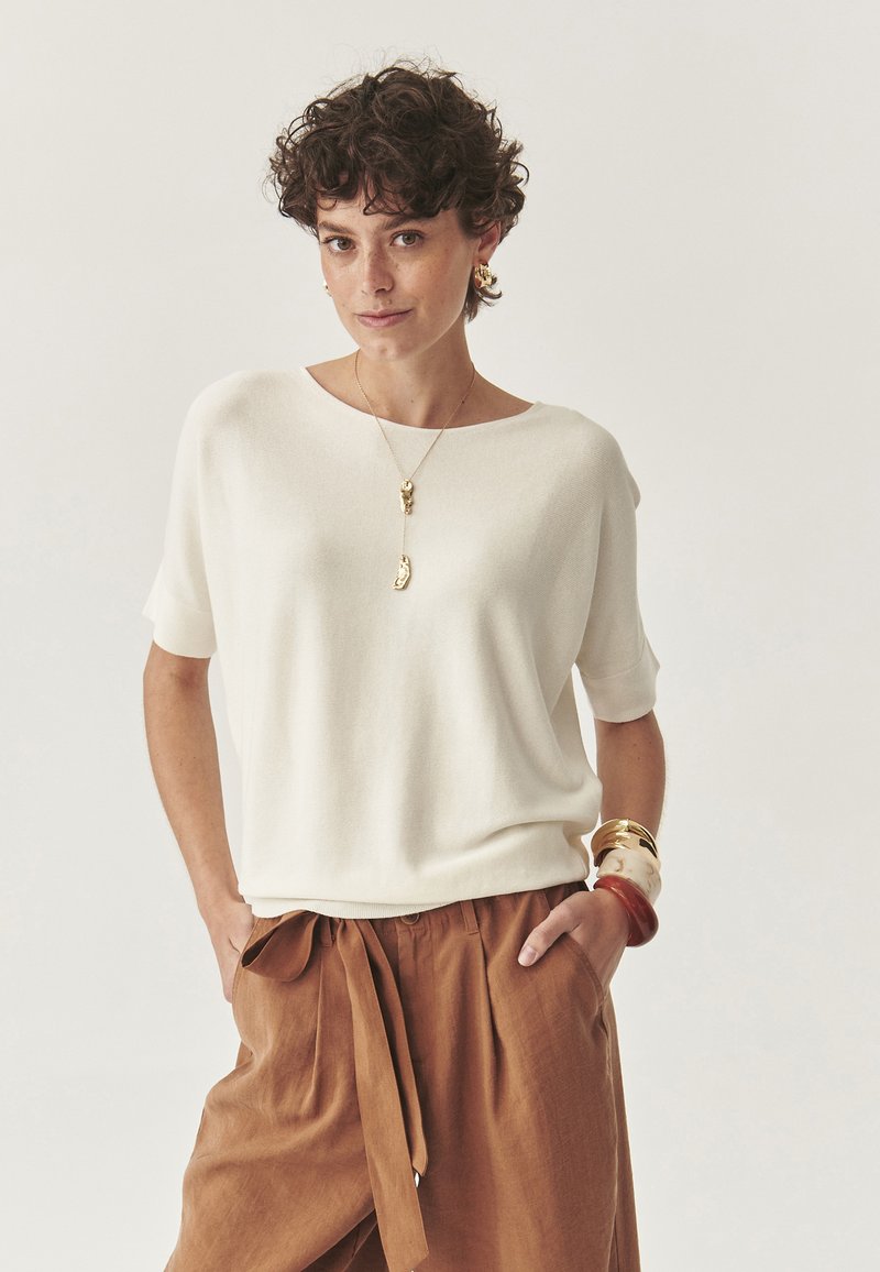 Letvægts, cremefarvet strikket sweater med en bred halsudskæring og korte ærmer, parret med brune, højtaljede bukser som er bundet i taljen.