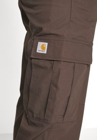 Pantalones cargo Carhartt marrones con un bolsillo con solapa y costuras visibles sobre la textura de la tela.