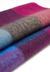 Opgevouwen wollen deken met een zachte textuur en een groot geruit patroon in roze, blauw, bruin en paarse tinten op een witte achtergrond.