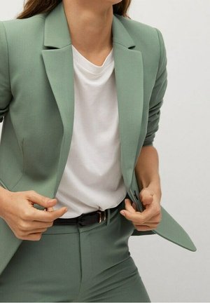 Persona con chaqueta y pantalones de traje verde claro y camiseta blanca metida por dentro, ajustando la parte delantera de la chaqueta con ambas manos.